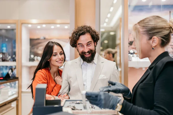 Gülümseyen ortağıyla birlikte heyecanlı bir adam tezgahta sergilenen pahalı saatlere bakarak perakende mağazasında satış asistanıyla etkileşime geçiyor.
