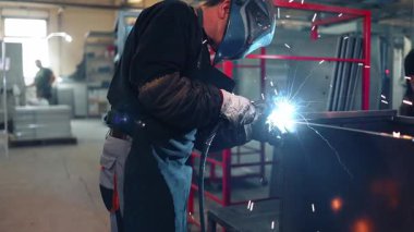 Koruyucu maske ve eldiven takan endüstriyel işçiler bir atölyede metal bir yapıyı kaynak yaparak yüksek sıcaklıktaki yay sürecinden parlak ışıklar ve kıvılcımlar yaratıyor.