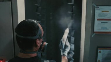 Solunum maskesi takan profesyonel bir erkek işçi. Endüstriyel sprey kabinindeki bir konveyör hattından sarkan metal parçalarına toz kaplaması uygulamak için sprey tabanca kullanıyor.