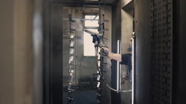 Solunum maskesi takan profesyonel bir erkek işçi. Endüstriyel sprey kabinindeki bir konveyör hattından sarkan metal parçalarına toz kaplaması uygulamak için sprey tabanca kullanıyor.