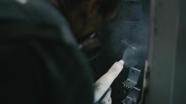 Solunum maskesi takan profesyonel bir erkek işçi. Endüstriyel sprey kabinindeki bir konveyör hattından sarkan metal parçalarına toz kaplaması uygulamak için sprey tabanca kullanıyor.