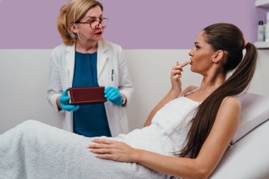 Mavi eldivenli ve laboratuvar önlüklü kozmetolog modern bir güzellik kliniğinde tedavi yatağında yatan gülümseyen bir kadın müşteriye çeşitli cilt ürünleri sunuyor.