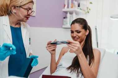 Mavi eldivenli ve laboratuvar önlüklü kozmetolog modern bir güzellik kliniğinde tedavi yatağında yatan gülümseyen bir kadın müşteriye çeşitli cilt ürünleri sunuyor.