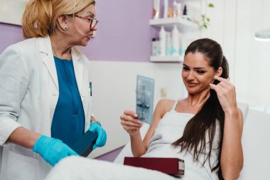 Kadın hasta şırıngayı incelerken, cildiyeci modern bir klinikte yüz dolgu seçeneklerini, güven, profesyonel bakım ve gençleşme beklentilerini açıklar.