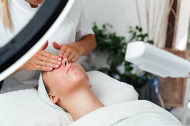 Güzel, rahat, genç bir kadın gözleri kapalı uzanmış güzellik merkezinde ya da kuaförde yüz masajı yapıyor. Bayan müşteri cilt bakımı tedavisinden zevk alıyor. Sağlık ve güzellik kavramı.