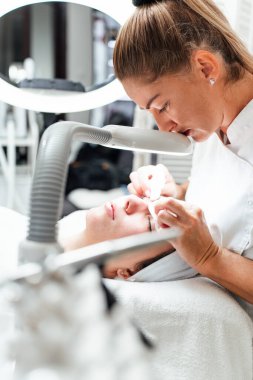 Profesyonel güzellik uzmanı, yüz estetiği ameliyatı sırasında kağıt mendille tedavi masasında yatan genç bir kadının burnundaki sivilceyi çıkarıyor.