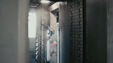 Solunum maskesi takan profesyonel bir erkek işçi. Endüstriyel sprey kabinindeki bir konveyör hattından sarkan metal parçalarına toz kaplaması uygulamak için sprey tabanca kullanıyor.