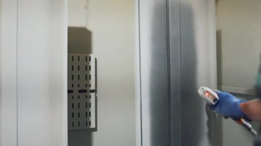 Solunum maskesi takan profesyonel bir erkek işçi. Endüstriyel sprey kabinindeki bir konveyör hattından sarkan metal parçalarına toz kaplaması uygulamak için sprey tabanca kullanıyor.
