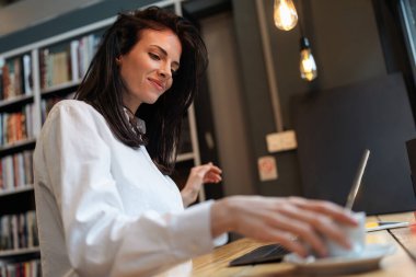 Beyaz gömlekli bir kadının, kahve masasında dizüstü bilgisayar kullanırken, kahve içerken, arka planda çalışma için sıcak asılı ışıklar ve kitaplık kullanırken yan görüntüsü..