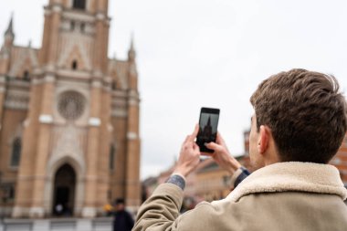 Bir adam arka planda tarihi bir şehir binası olan arka plan manzaralı bir selfie çekmek için akıllı telefon tutuyor..