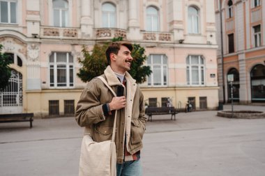 Gülümseyen adam dışarıda elinde bir akıllı telefon ve çantasıyla tarihi şehir binalarının önünde duruyor..