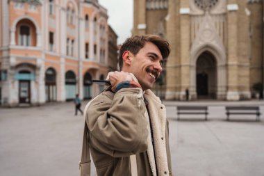 Gülümseyen genç bir adam arkasında tarihi kilise mimarisi olan bir Avrupa şehir meydanında yürürken sırtında bir çanta taşıyor..
