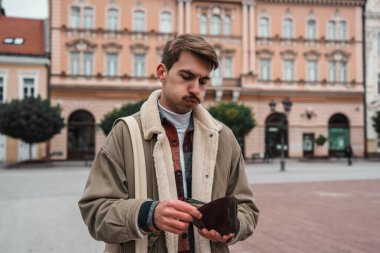 Endişeli bir genç adam arka planda tarihi binalar olan şehir meydanında dururken boş cüzdanına bakar..