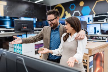 Gülümseyen genç bir çift, modern bir teknoloji mağazasında büyük bir düz ekran televizyona bakarak ev eğlencesi için alışveriş yapıyor..