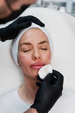 Siyah eldivenli profesyonel bir kozmetik uzmanı cilt bakımı sırasında bir kadının yüzünü temizler. Modern bir klinikte üst düzey estetik ilaç tedavisi.