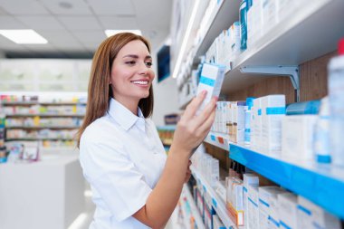 Beyaz önlüklü, gülümseyen bir kadın eczacı modern bir eczanede ilaç ve cilt ürünlerini düzenleyip kontrol ediyor..