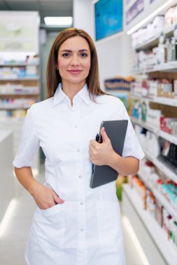 Beyaz önlüklü kendine güvenen bir kadın eczacı elinde dijital tabletle iyi aydınlatılmış bir eczanede duruyor. Modern sağlık hizmetleri, tıbbi uzmanlık ve ilaç hizmetlerini temsil ediyor..