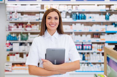 Beyaz laboratuvar önlüklü, dost canlısı bir kadın sağlık görevlisi, dolu bir eczanede rafların önünde dururken dijital tablet tutuyor. Gülümsüyor ve kameraya bakıyor..