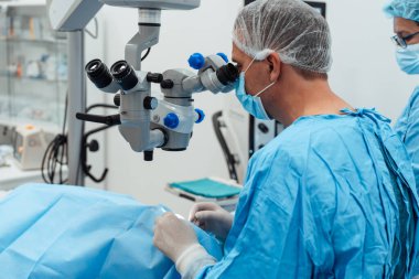 Uzman bir göz cerrahı steril bir ameliyathanede tıbbi personelle birlikte hassas bir ameliyat sırasında yüksek hassasiyetli bir mikroskop kullanır..