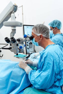 Uzman bir göz cerrahı steril bir ameliyathanede tıbbi personelle birlikte hassas bir ameliyat sırasında yüksek hassasiyetli bir mikroskop kullanır..