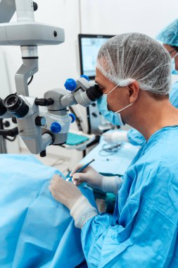 Uzman bir göz cerrahı steril bir ameliyathanede tıbbi personelle birlikte hassas bir ameliyat sırasında yüksek hassasiyetli bir mikroskop kullanır..