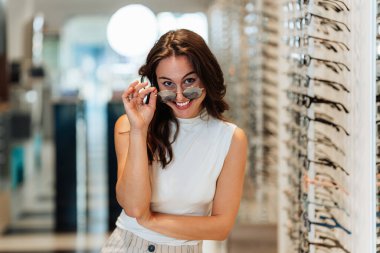 Neşeli genç bir kadın, güneş gözlüğünün üzerinden, parlak, modern bir gözlükçüden göz giysisi alırken, neşeyle bakar..
