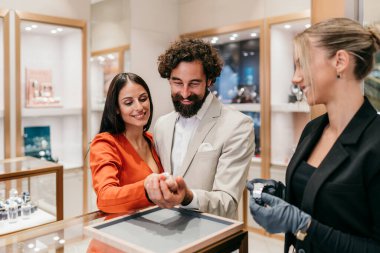 Sofistike bir çift, lüks bir butikte bir nişan yüzüğü ve lüks saatler inceliyor. Profesyonel bir satış asistanının yardımıyla sıcak, altın bir bataklıkta..