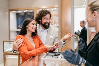 Gülümseyen bir çift lüks bir saat ya da mücevher seçerken, profesyonel bir satıcı onlara şık bir perakende mağazası ortamında yardımcı olur..