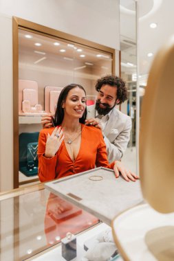 Lüks bir butikte aksesuar alışverişi yapan mutlu bir çift. Adam ortağına aynanın önünde zarif bir altın kolye denemesinde yardımcı olur. O da aynadaki yansımasına gülümser..