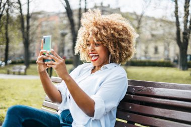 Sarışın, Afro saçlı heyecanlı bir kadın bankta oturmuş video görüşmesi yapıyor ya da parkta selfie çekiyor..