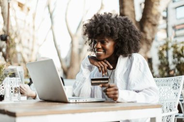 Gülümseyen Afro-Amerikalı bir kadın açık bir kafede otururken kredi kartı ve dizüstü bilgisayar kullanarak online alışveriş yapıyor..