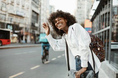 Afro saçlı neşeli bir Afro-Amerikalı kadın, bir arkadaşını selamlamak için elini sallıyor ya da sırt çantası ve laptop çantasıyla kalabalık bir şehir ortamında gidip gelirken..