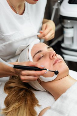 Profesyonel bir estetisyen modern bir güzellik kliniğinde rahatlamış bir kadın hasta üzerinde yüz yenileme işlemi için özel bir tıbbi cihaz kullanır..