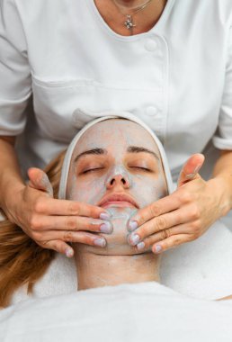 Profesyonel bir güzellik uzmanı, bir sağlık merkezinde cilt bakımı sırasında rahatlamış bir kadının yüzüne mineral çamur maskesi uygular..