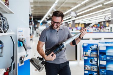 Neşeli bir erkek perakende satış elemanı, kablosuz elektrik süpürgesi modern bir ev aletleri galerisinde elektro gitarmış gibi davranarak çalışırken eğleniyor..