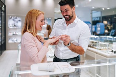 Gülümseyen bir adam modern bir kuyumcuda bir kadının parmağına nişan yüzüğü takıyor. Çift, lüks aksesuarlar alırken romantik bir kilometre taşını kutluyor..