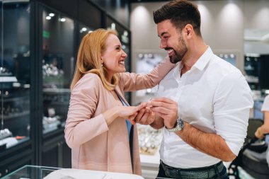 Gülümseyen bir adam modern bir kuyumcuda bir kadının parmağına nişan yüzüğü takıyor. Çift, lüks aksesuarlar alırken romantik bir kilometre taşını kutluyor..