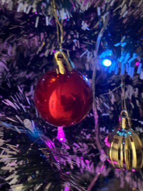 Süslü bir ağaçtan sarkan parlak kırmızı bir Noel süsü. Arka plandaki renkli ışıklar tatil sezonunun neşesini ve kutlamasını yansıtan neşeli bir atmosfer yaratıyor..