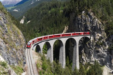Kızıl bir yolcu treni, İsviçre Alplerinde bir mühendislik harikası olan ikonik Landwasser Viaduct 'tan geçiyor ve Rhaetian Demiryolu' nun güzelliğini ve çarpıcı dağ çevresini gözler önüne seriyor..