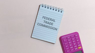 Bir spiral not defterine yazılmış federal ticaret komisyonu canlı mor bir hesap makinesinin yanında oturuyor. Bu da tüketici korumasını veya hafif bir yüzey üzerindeki ticari uyum analizini öneriyor..