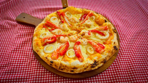 A freshly baked seafood pizza served on a wooden paddle board, placed over a red and white checkered tablecloth. The pizza is topped with shrimp, squid rings, and imitation crab sticks, along