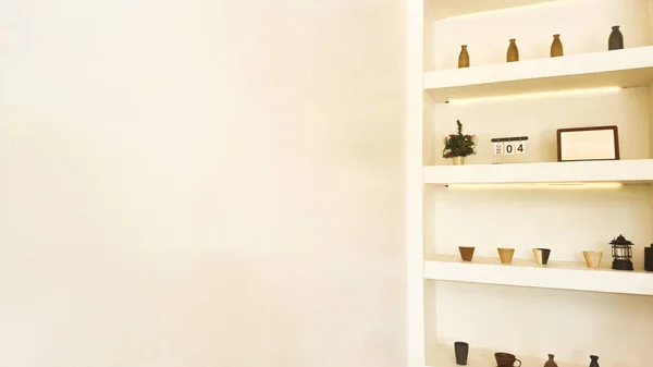 A bright minimalist interior featuring built-in white shelves decorated with simple pottery, small vases, a framed artwork, a tabletop calendar, and a small ornamental plant. Warm lighting highlights
