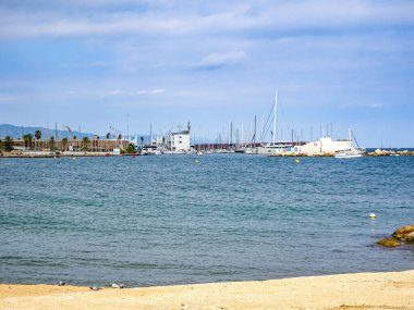 Barselona 'da. İspanya. 2 Ekim 2025. Port Olimpic yat limanının sakin deniz manzarası, yelkenli yatları ve açık gökyüzü. Huzurlu kentsel kıyı manzarası ön planda kumlu plaj.