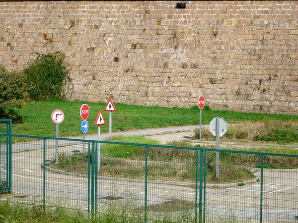 group of road signs near a stone fortress wall and green fence in area. The signs include STOP, yield, and directional symbols.