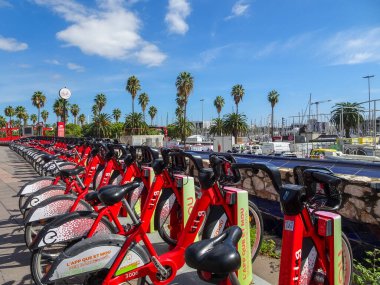 Barselona, İspanya. 6 Ekim 2025. Port Vell yakınlarında, güneşli bir gökyüzünün altında, palmiye ağaçları olan bir bisiklet paylaşım istasyonuna park edilmiş kırmızı bisiklet kuyruğu..