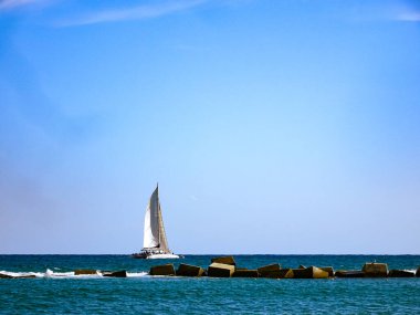 Akdeniz boyunca açık mavi bir gökyüzünün altında dalgakıran yakınında beyaz bir yelkenli yelken açar.