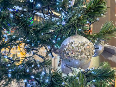 Yeşil iğneler ve şenlikli mavi ışıklarla çevrili ağaç dalında pullarla parıldayan gümüş Noel süsü..