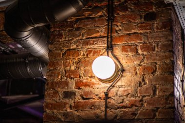 Endüstriyel boruları ve yuvarlak bir aydınlatma armatürü olan eski bir tuğla duvara yakın çekim yaparak kırsal bir kentsel atmosfer yaratıyor..