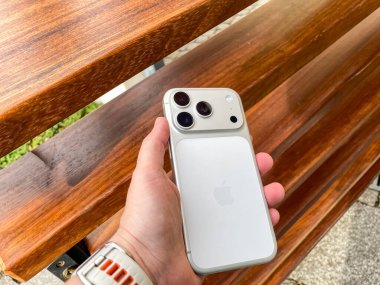 Yılan pençesi. Polonya. 14 Kasım 2025. Açık havada ahşap bir bankta MagSafe bataryası olan gümüş bir iPhone 'u tutan bir el. Telefonun parlak tasarımını aydınlatan doğal bir ışık..