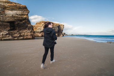 Galiçya, İspanya 'daki Las Katedrallerin kumlarında koşan bir kadın uçurum oluşumları ve okyanus hareketlerini gösteriyor ve dinamik bir seyahat ve keşif sahnesi yaratıyor.